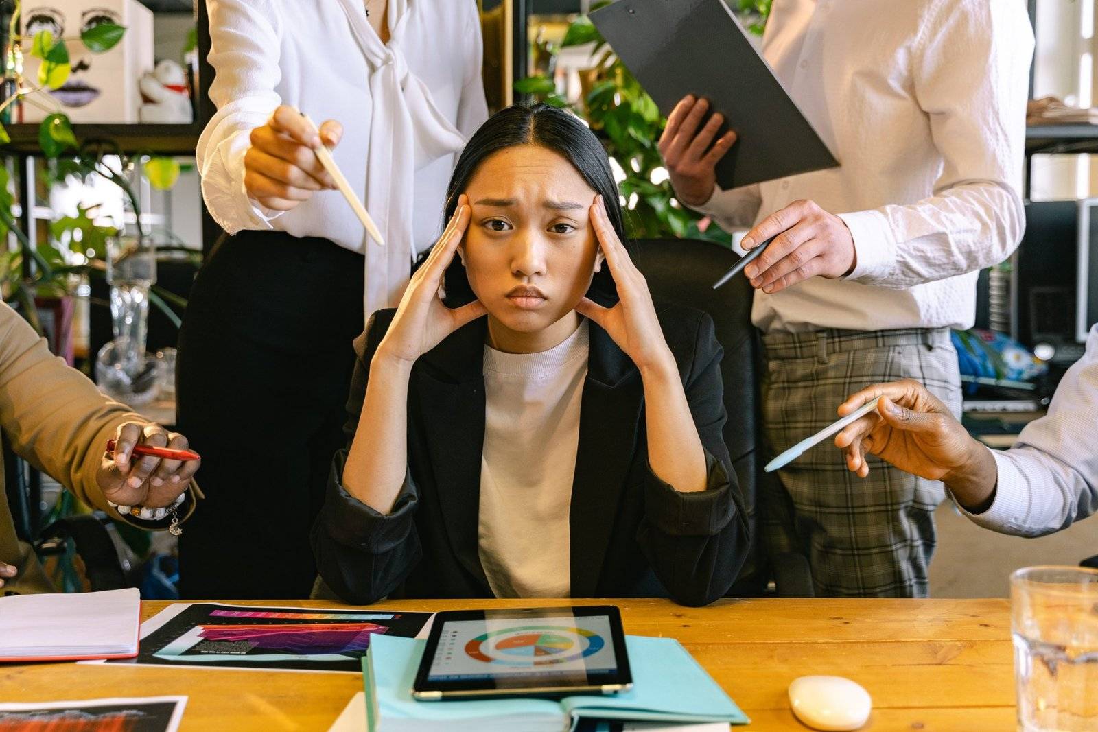 overwhelmed woman sitting at desk with people surrounding her in the workplace suffering from hustle culture syndrome
